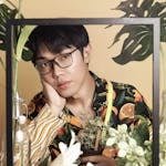 Fashionable male model with eyeglasses posing among vibrant plants in an indoor setting.